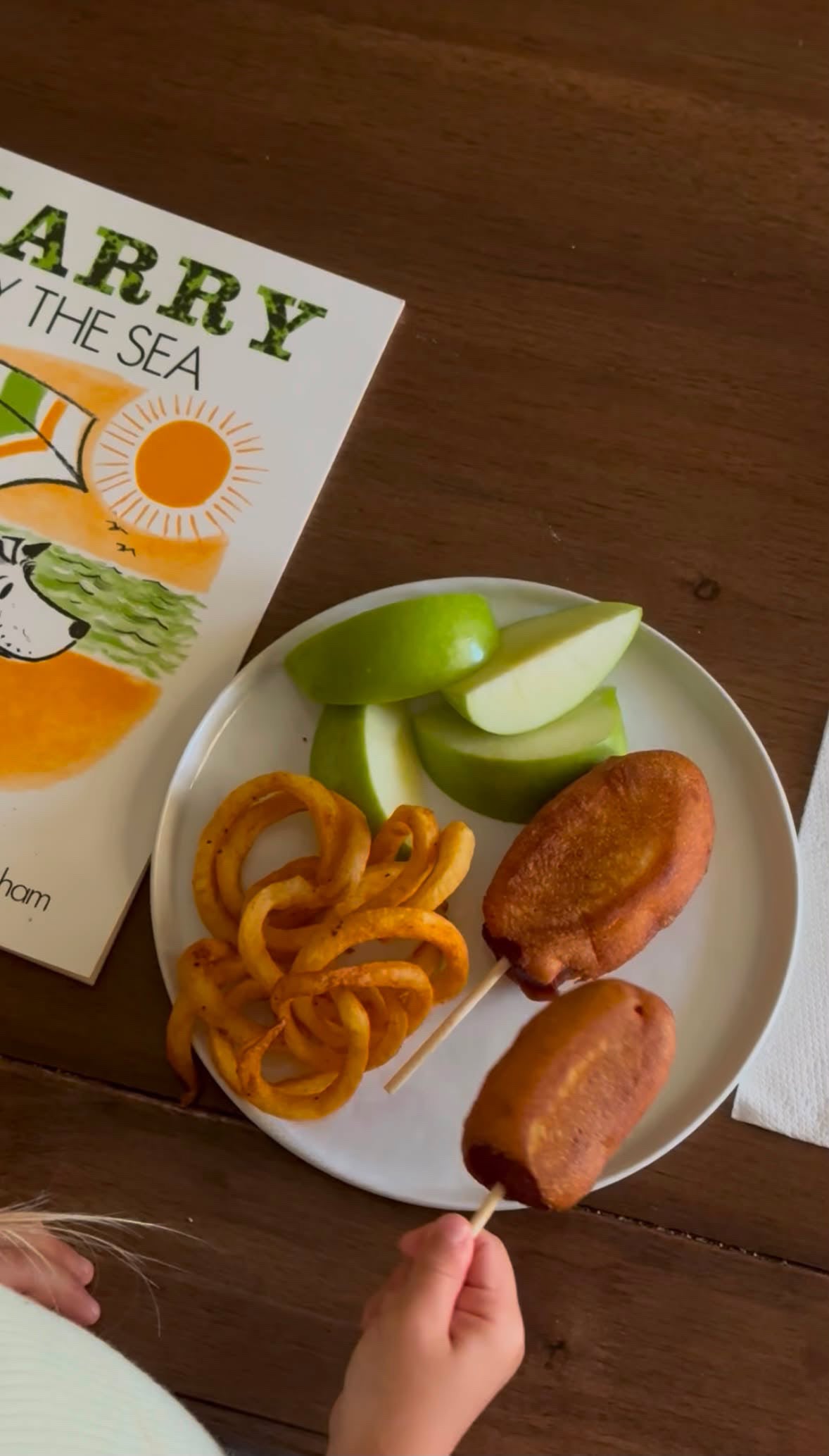 A child's hand holds a corn dog on a wooden table next to a plate with apple slices, curly fries, and two fried fish cakes, with a children's book titled 'Harry the Sea' nearby.