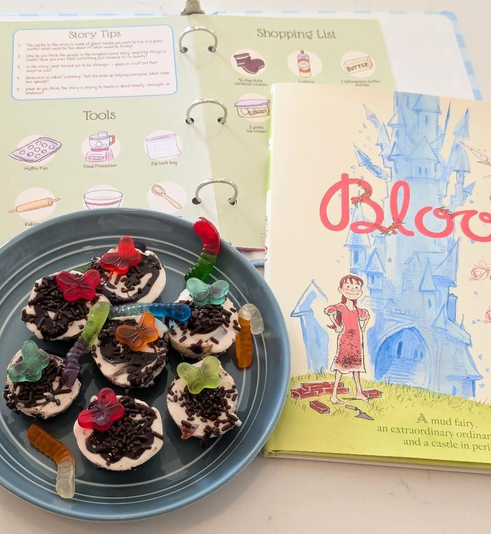 A styled flat-lay of a children's picture book titled 'Bloom' next to a blue plate of mini chocolate cake treats topped with gummy worms, with an open ring cook book showing story tips and shopping list pages.