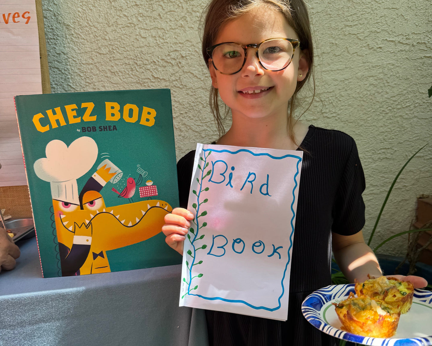 Young girl holding a book titled 'Chez Bob' and a sign reading 'Bird Book' with a plate of food.