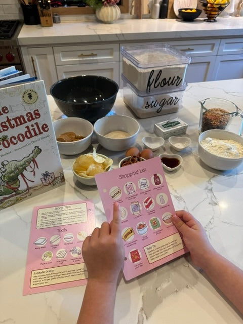 A person's hands holding a pink 'Shopping list' recipe book in a bright kitchen, surrounded by baking ingredients on a marble countertop, including bowls of flour, sugar, butter, eggs, and a 'The Christmas Crocodile' book.