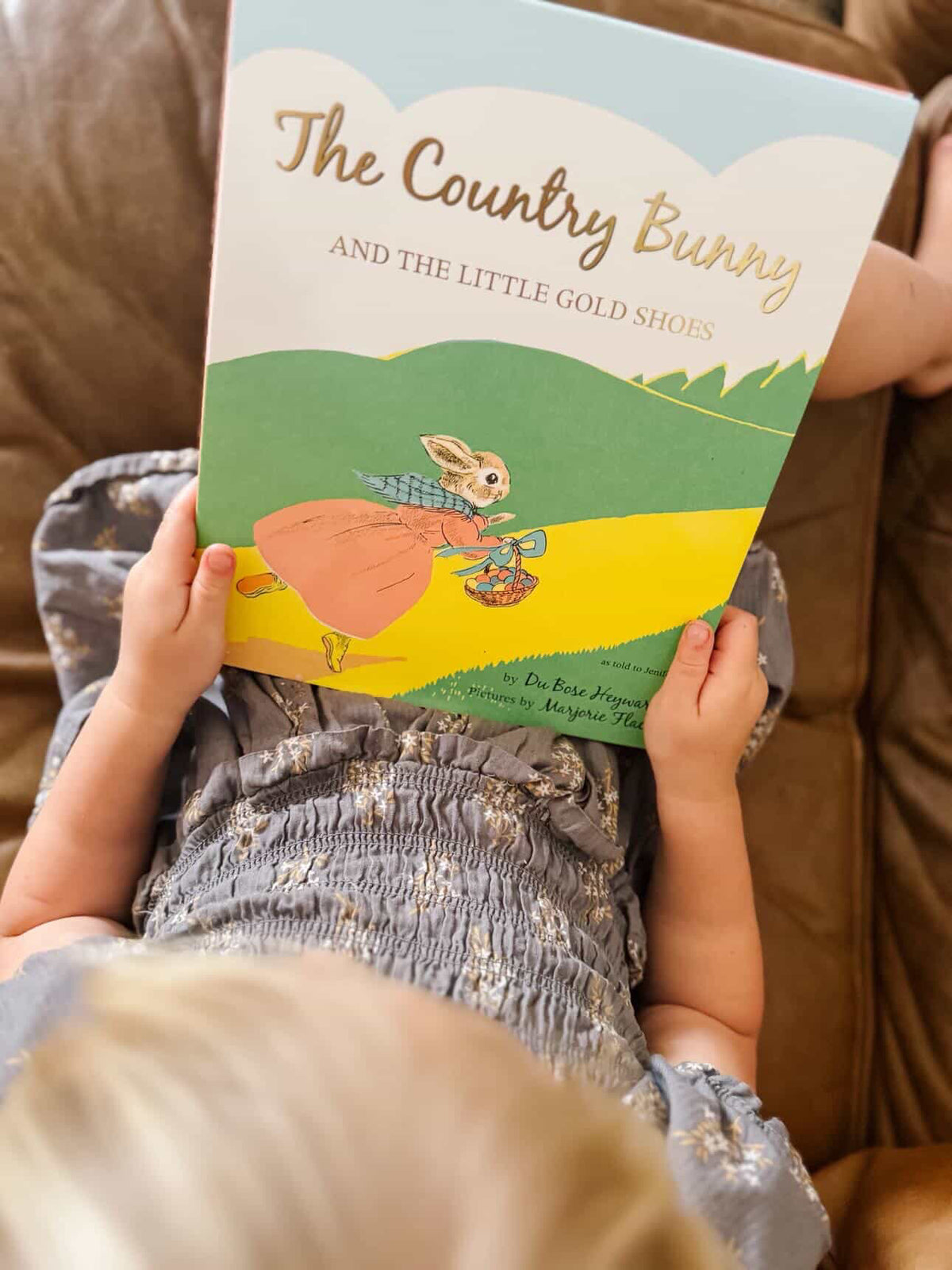 A child holds a children's book titled "The Country Bunny and the Little Gold Shoes" while lying on a brown couch. The book cover shows a bunny in a pink dress on a yellow path with green hills under a light blue sky.