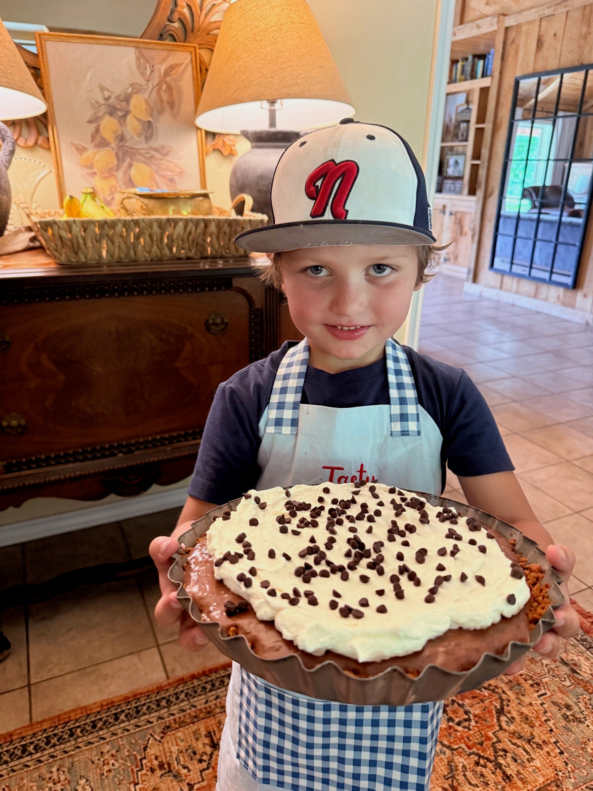 A young boy wearing a navy and white baseball cap with a red 'N' logo, a navy t-shirt, and a blue-and-white gingham apron holds a freshly baked chocolate pie topped with white whipped creram and chocolate chips in a scalloped metal pie dish, set against a warm, rustic indoor background.