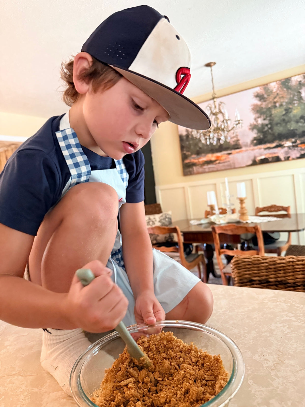 A young boy wearing a navy and white baseball cap, a navy t-shirt, and a blue-and-white gingham apron kneads a brown graham cracker crumb mixture in a glass bowl on a counter top in a home setting.