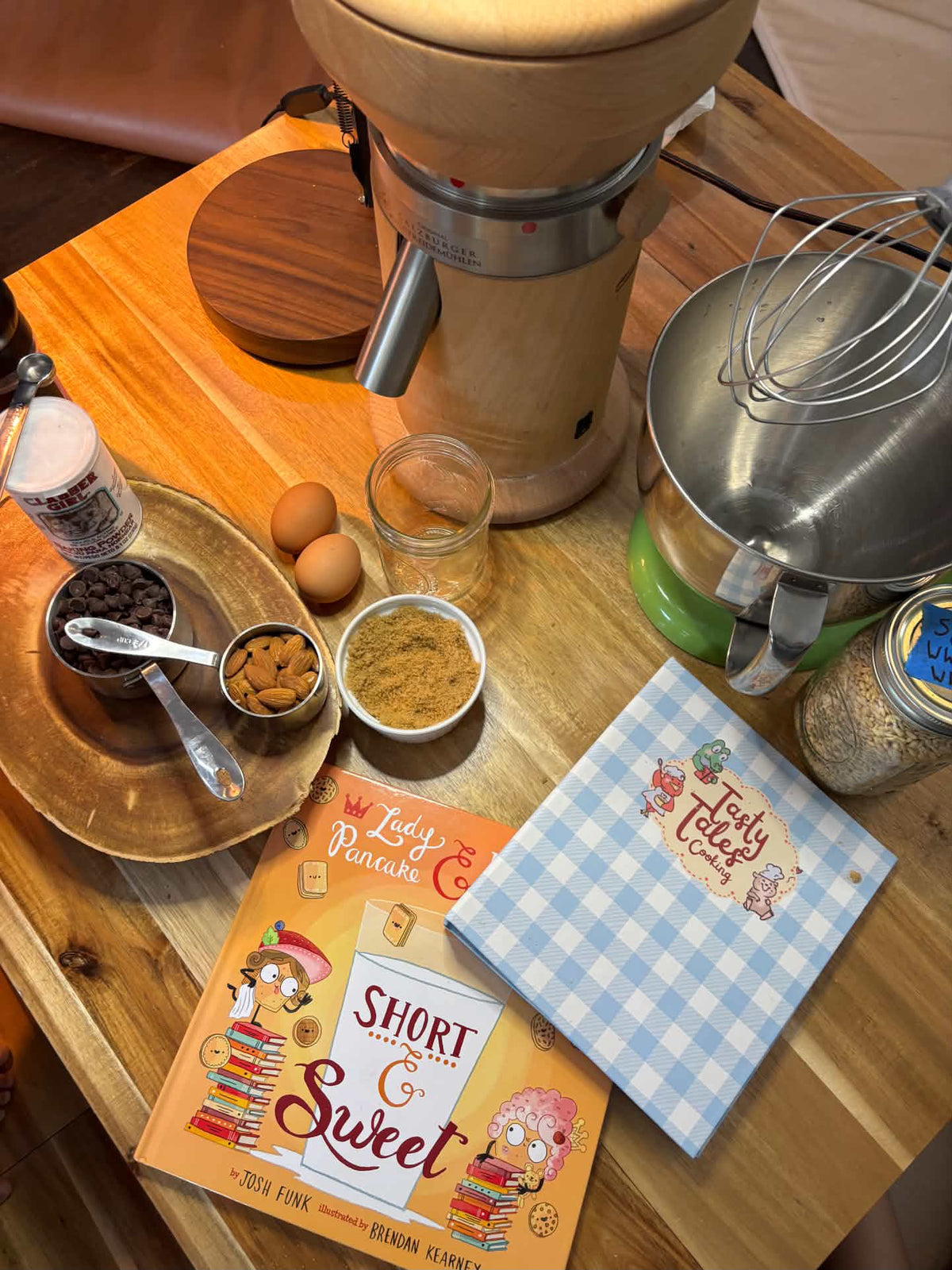 A wooden countertop setup with baking tools and baking ingredients, including a stand mixer, a whisk, bowls of cocoa powder and spices, eggs, a blue and white checkered Tasty Tales cookbook, and a picture book  titled 'Short & Sweet' by Josh Funk. 