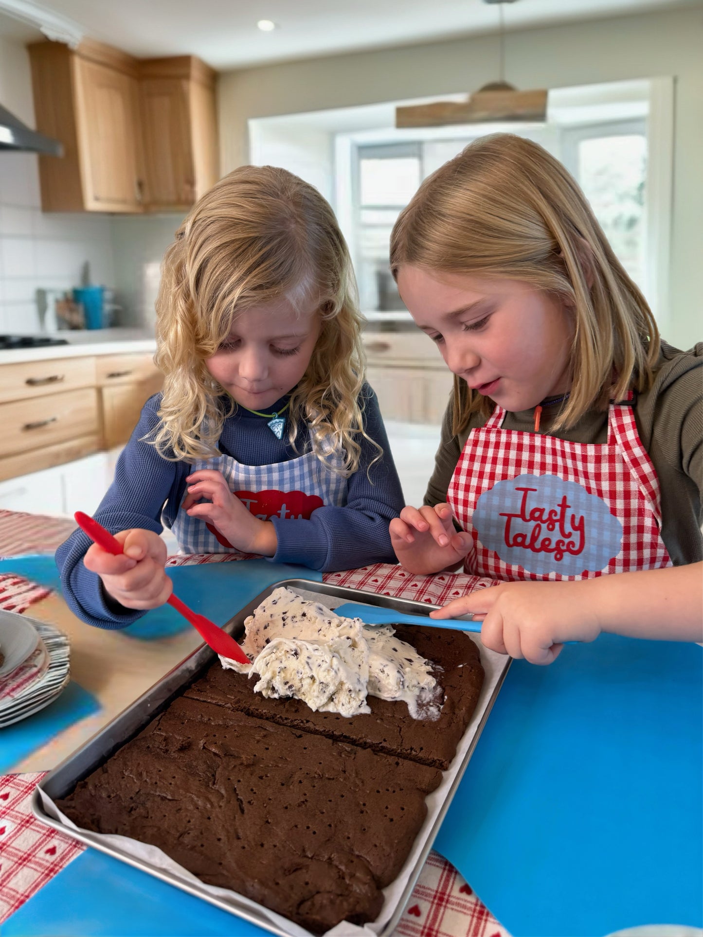 Two children in a kitchen, one wearing an apron with 'Tasty Tales' text, preparing a dessert.