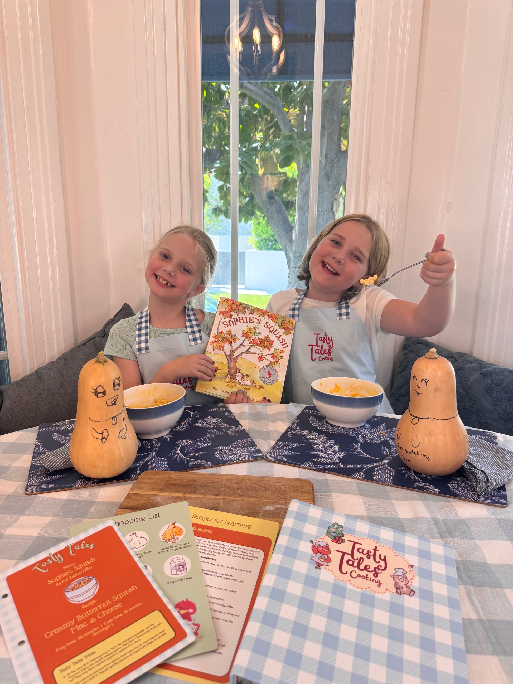 Two girls showing off their butternut squash mac and cheese creation at a kitchen table.  One girl is holding a copy of Sophie's Squash.  In the foreground are Tasty Tales recipe cards and cookbook binder.